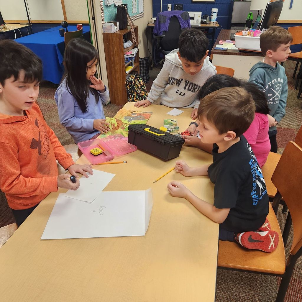 Several students lean over a table studying clue papers, a toolbox, and a pink supply box as they collaborate to solve the breakout challenge.
