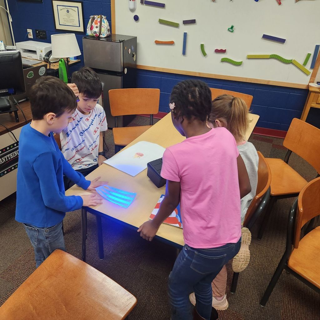 Four students shine a blue UV flashlight onto a clue sheet on the table. They are working together near a toolbox and a book from the activity.