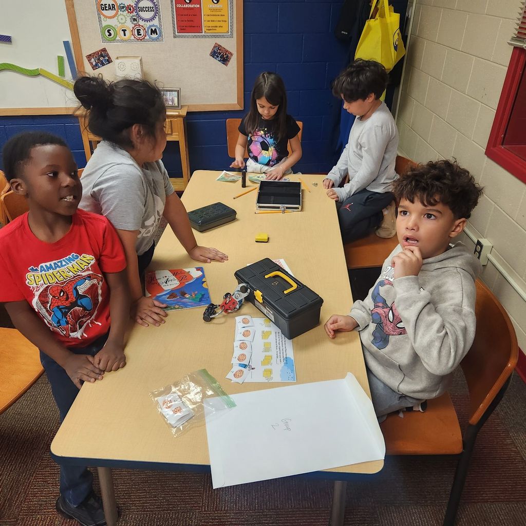 Five students gather around a table with lock boxes, clue cards, and a copy of How to Make an Apple Pie and See the World. One student smiles at the camera while others focus on solving the puzzle.