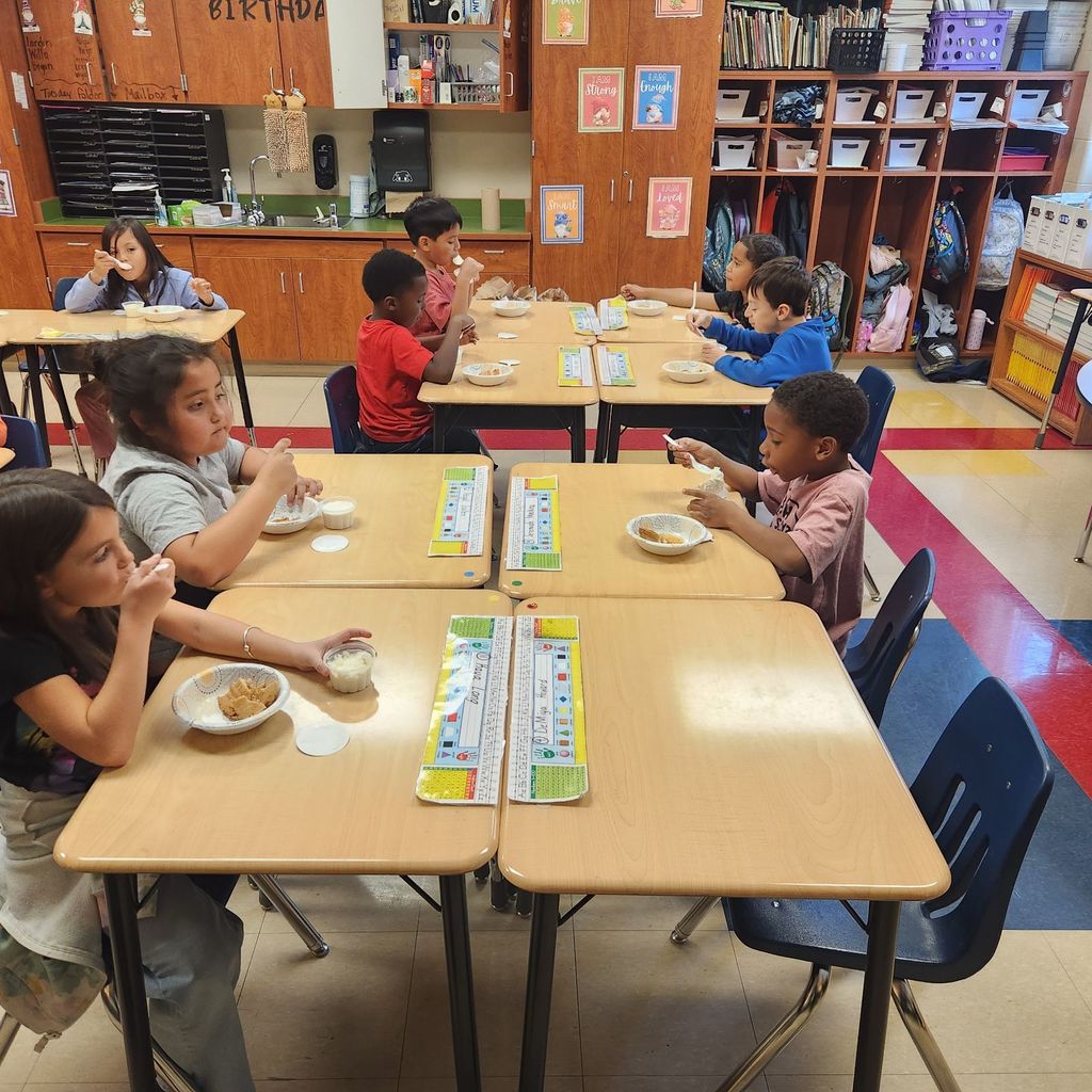 Students sit at their desks in the classroom enjoying bowls of apple pie and vanilla ice cream. Each child has a small cup and spoon as they celebrate after the activity.