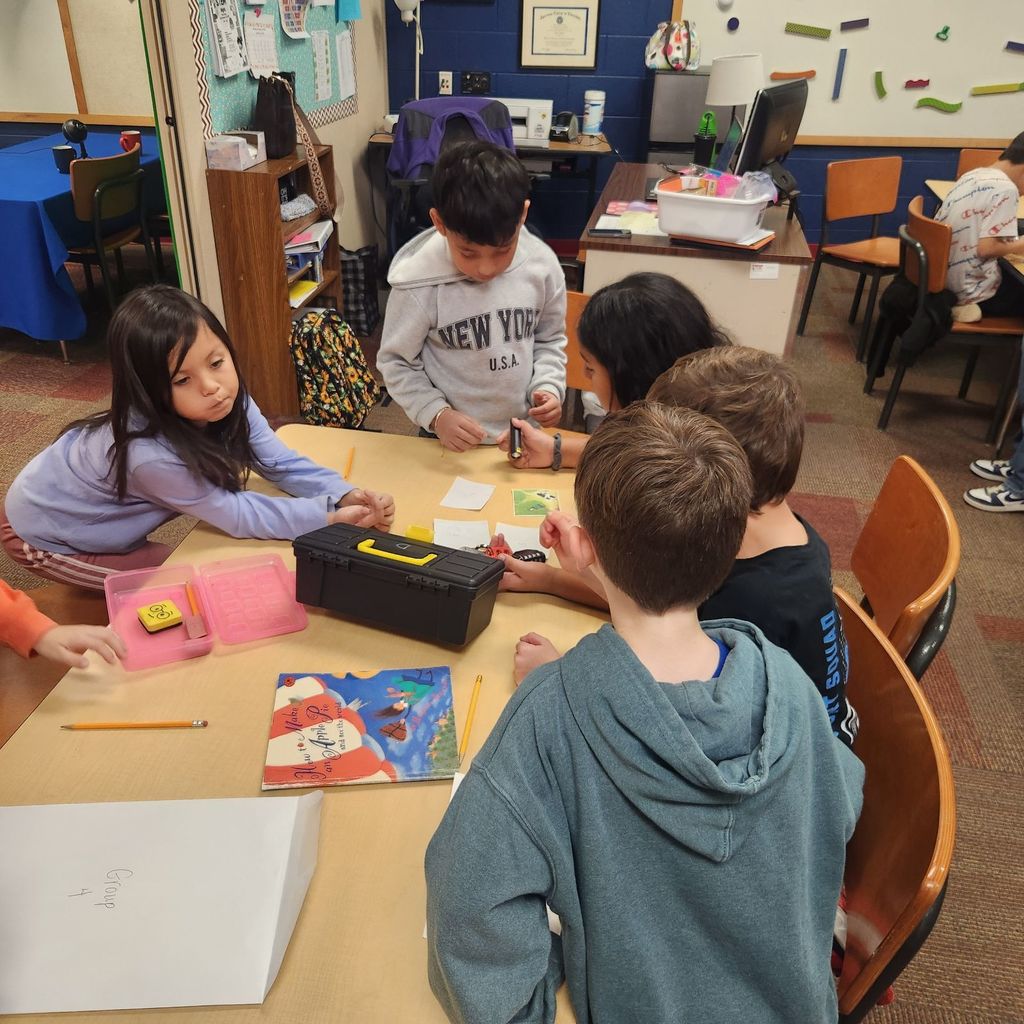 A group of young students sit and stand around a table working together on Breakout EDU clues. One student holds a small flashlight while others look at papers, a toolbox, and the book How to Make an Apple Pie and See the World spread out on the table.