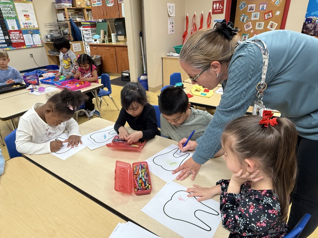 Several young students sit at a table coloring large tooth-shaped outlines on paper. A teacher leans over to help one student while others focus on their work. Crayons and supply boxes are scattered on the table.