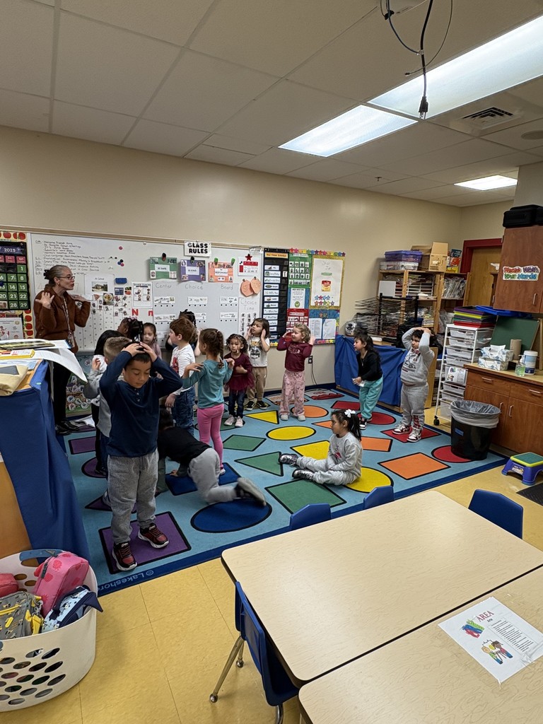 A classroom full of preschool students stands and moves around on a colorful carpet during an activity. A teacher stands to the left giving instructions while some children follow motions and others sit or move around the carpet.