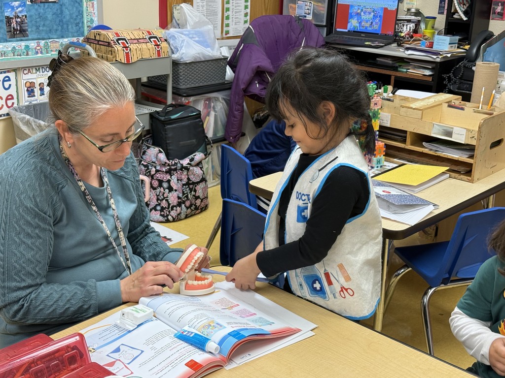 A teacher sits at a small table with a young student who is wearing a pretend doctor vest. The teacher holds a model of teeth while the student points at it, practicing how to brush. An open health book, toothbrush, and school supplies are on the table.