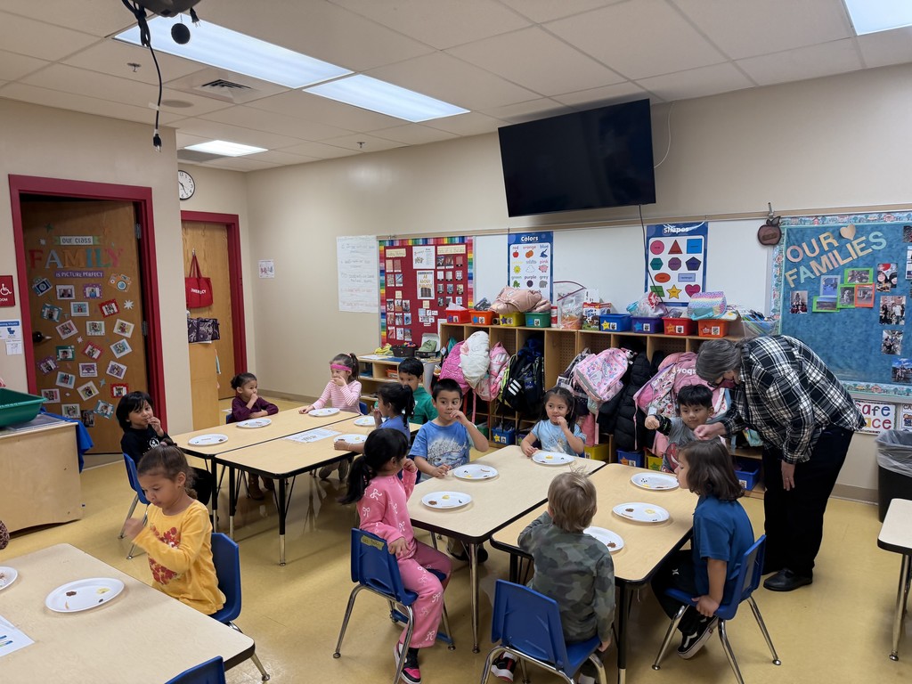 A preschool classroom with children sitting at tables, each with a paper plate of small food items for a tasting activity. A teacher stands beside one table assisting a student. Backpacks and coats hang along the cubbies in the background.