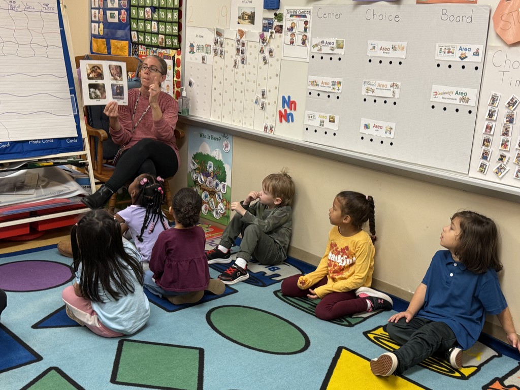 A teacher sits in a rocking chair at the front of the classroom holding up a picture book and speaking to a group of young children sitting on a colorful carpet. The children watch and listen attentively.