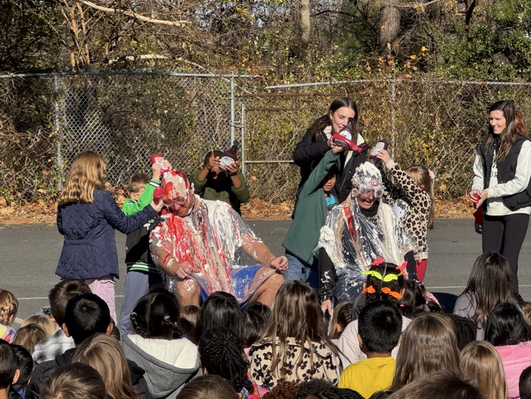 A group of students watching two adult adults, get covered in strawberry and chocolate syrup