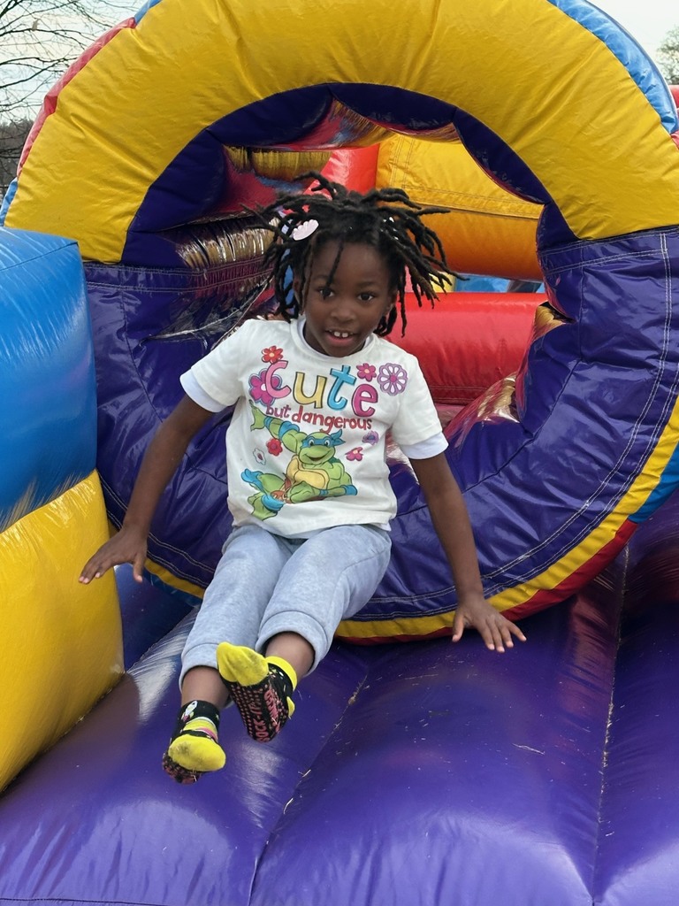 A young girl slides out of a bright yellow, purple, and red inflatable tunnel. She has braids and is smiling as she leans out onto the inflatable surface.