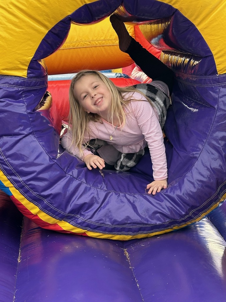A girl with long blond hair is crawling through a circular inflatable tunnel. She is smiling and partially upside down inside the colorful bounce-house obstacle.