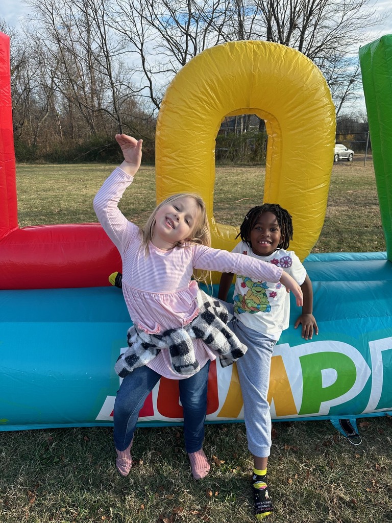 Two children pose happily in front of a large inflatable structure with big colorful letters. One stands behind the letter “O,” and both children have their arms outstretched and are smiling.