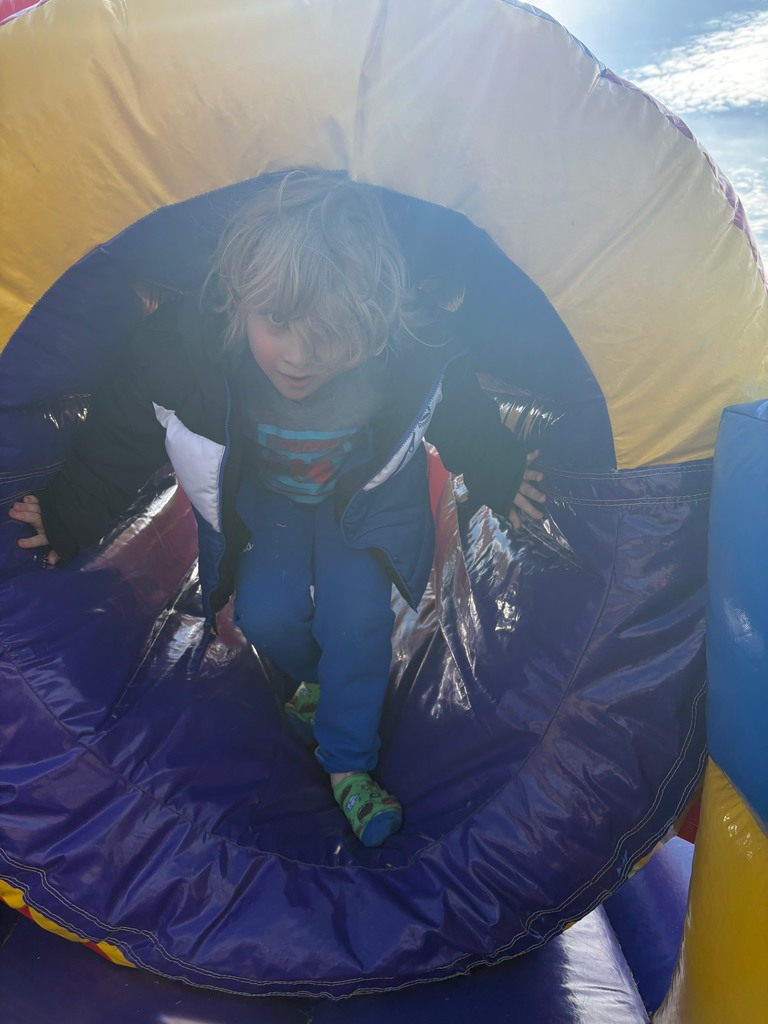 A child with shaggy blond hair climbs through a purple and yellow inflatable tunnel. The sun creates a bright glow in the top corner of the image.