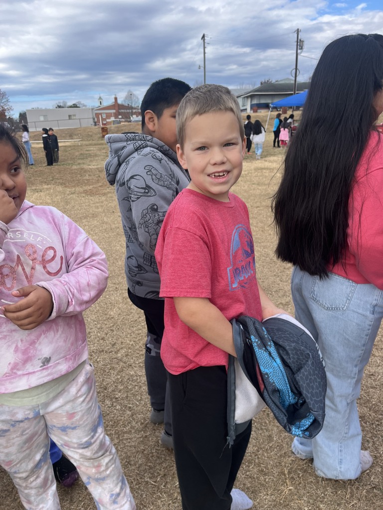 A young boy in a red T-shirt stands outside in a line with other children. He is smiling and holding a jacket. The group is on a grassy field with other students in the background.