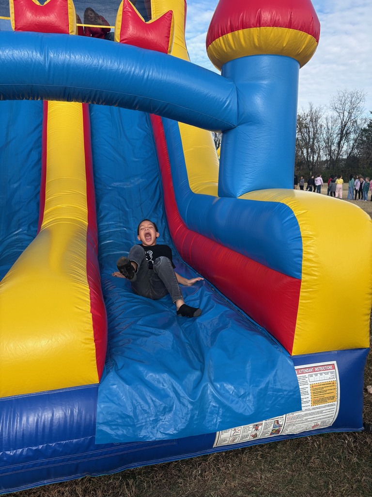 A boy is sliding down a large inflatable slide with blue, red, and yellow sections. He has a big open-mouthed smile as he reaches the bottom.