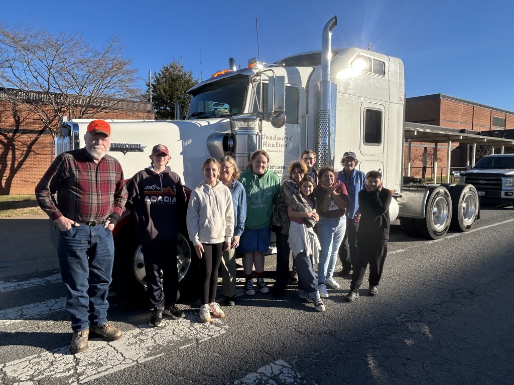 This is a daylight photograph of a diverse group of approximately ten individuals, including adults and children, posing together in front of a large, silver semi-truck. The semi-truck, which appears to be a tractor unit with "Deadwood Hauling" visible in green lettering on the driver's side door, is parked on what looks like a street or parking lot with a crosswalk. The group is standing closely together in front of the truck's grille and wheels. On the far left, an older man with a white beard wearing a plaid shirt, blue jeans, work boots, and an orange cap is standing with his hands on his hips. The rest of the group includes various ages, wearing casual clothing such as sweatshirts and jeans. The background consists of a brick building to the right, possibly a school or commercial building, and some bare trees to the left, suggesting a cooler season. The sky is bright blue, and the sun is shining brightly from behind the truck on the right, causing a strong lens flare near the chimney/exhaust stack.