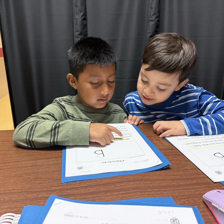 two youn male students sit at a table and look over a book. other papers and items are on the table as well 