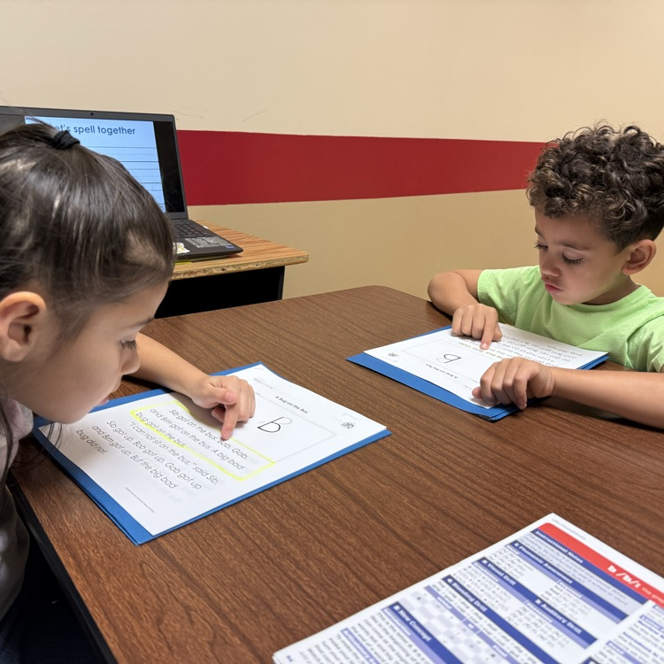 two young students sit across a table from one another and read site words off of a sheet of paper laid in front of them  