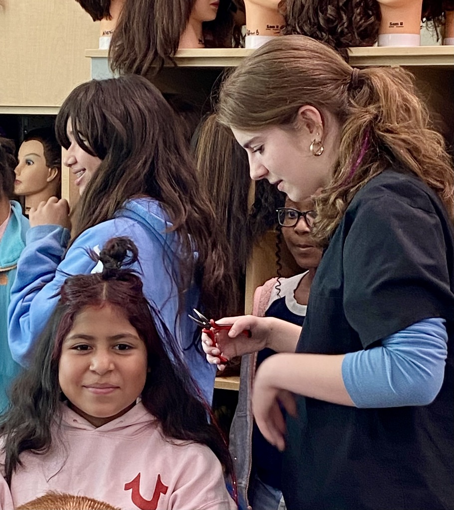 Two students practice hair in a cosmetology classroom with a student helping.  Another student looks on with a smile.