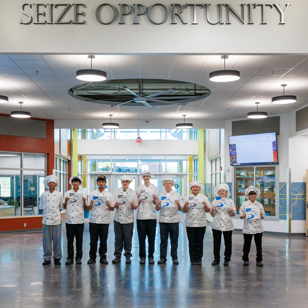 Eleven culinary students wearing white chef coats, hats, and black pants stand in a row inside the CTEC lobby beneath the large “SEIZE OPPORTUNITY” sign. Each student holds a certificate, smiling proudly for the photo. The bright, modern lobby and overhead lighting create a clean, professional backdrop.