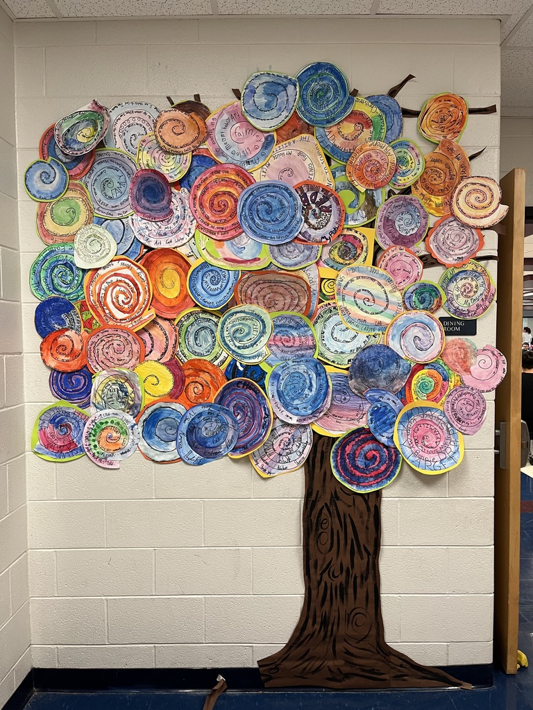 A large, colorful mural of a tree displayed on a school hallway wall. The tree trunk and branches are made from brown paper, while the treetop is composed of many circular, spiral-patterned paper artworks created by students. Each spiral is brightly painted in various colors—pink, blue, green, orange, yellow, and purple—and many include handwritten messages or names around the spirals. The artwork forms a full, vibrant canopy that covers most of the wall space.