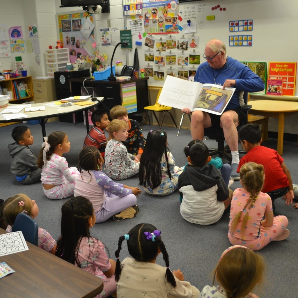 Teacher sits in front of a kindergarten class and reads a book in the classroom.