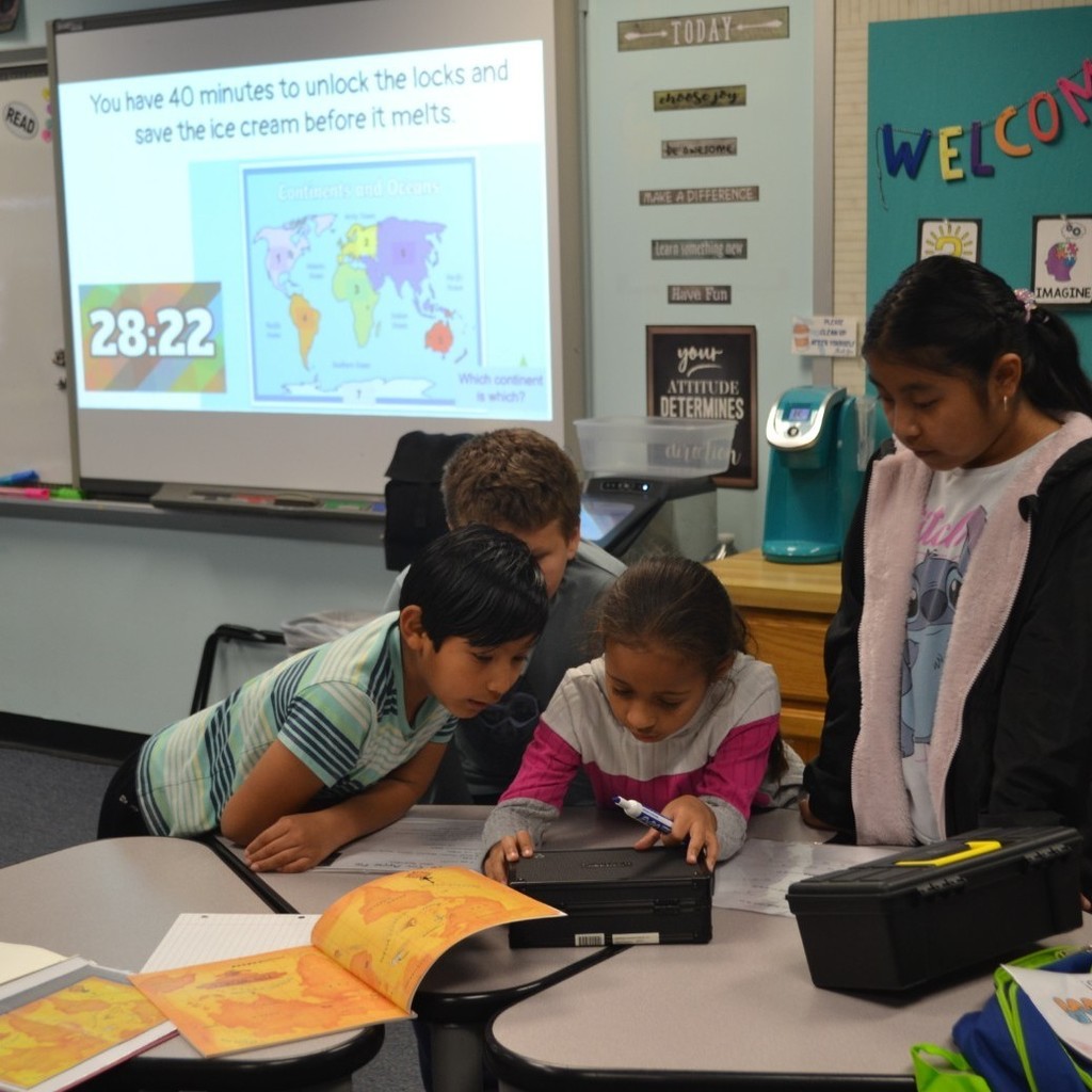 Four students work together solving clues in a classroom to open lock boxes. Map of continents on the board.