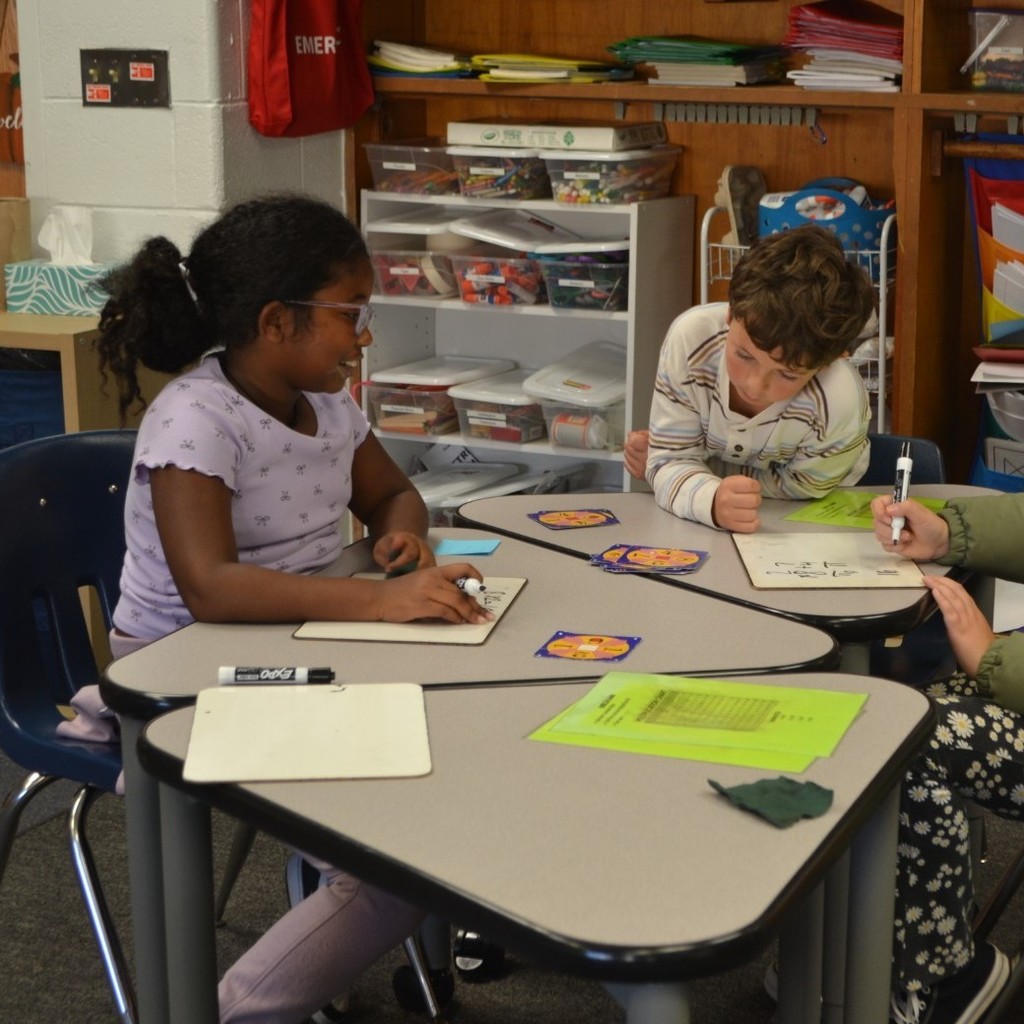 Two girls and a boy play Math 24 in their classroom while eating lunch.
