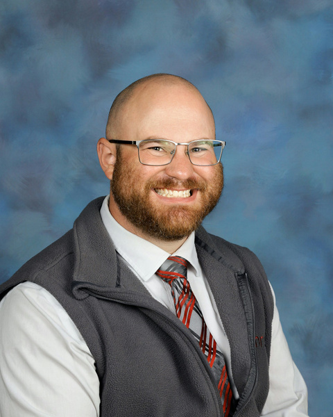 A smiling man with glasses and a beard, wearing a gray vest over a white shirt and a red-striped tie. The background is a soft, blurred blue.