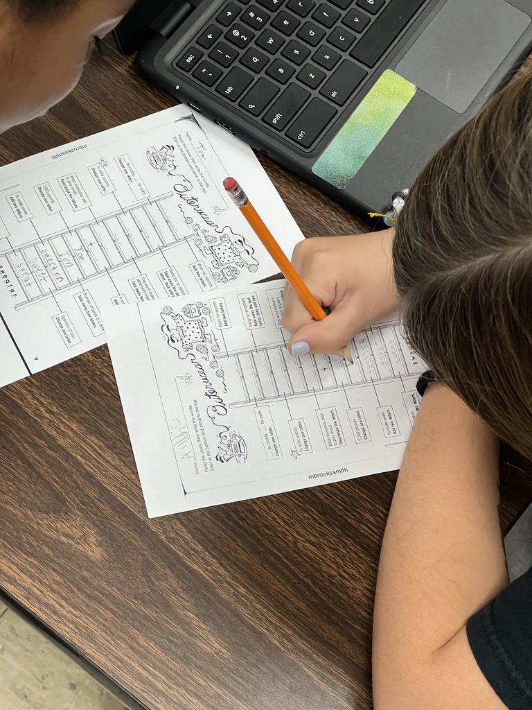 Two students are working together on printed “Outerwear” vocabulary worksheets. Each student is writing answers in pencil while sitting at a desk with a Chromebook nearby.