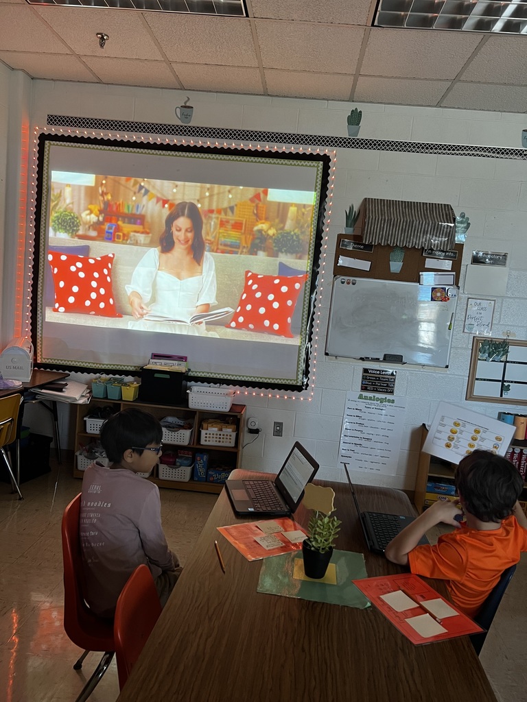 A classroom scene where a projector displays Lea Michele reading Rosie Revere, Engineer. Two students sit at a table with Chromebooks and bright orange theme-finding worksheets. A small plant is on the table, and classroom decor surrounds the projection screen.