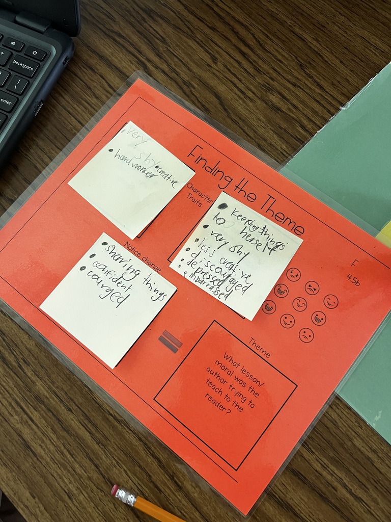A laminated orange worksheet titled “Finding the Theme” sits on a wooden table. Several sticky notes with handwritten character traits and observations are placed on it. A pencil rests nearby, and part of a Chromebook keyboard is visible on the left.