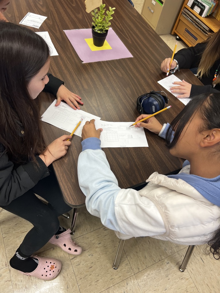 Two students sit at a table working together on logic puzzle worksheets. One student is pointing at the paper while the other writes with a pencil. A small potted plant decorates the middle of the table.