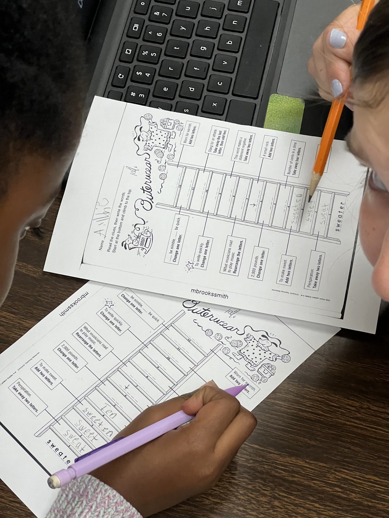 Close-up of students’ hands working on math logic puzzle sheets. One worksheet shows boxed grids with numbers and variables, and another sheet is placed above it on the desk.