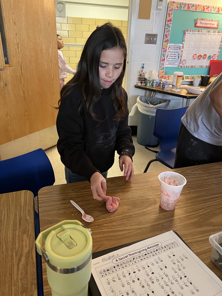 A student kneads or stretches pink slime at a table. A nearly empty plastic cup of slime ingredients sits nearby. Another student and classroom decorations are visible in the background. A worksheet titled “A Secret Thanksgiving Message” is on the table.