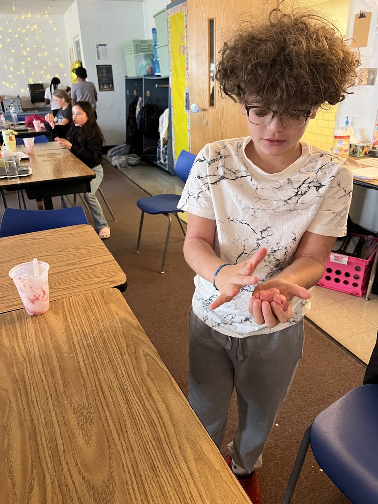 A student with curly hair and glasses stands at a table shaping a ball of pink slime in their hands. Other students are working around the classroom in the background. A plastic cup with slime residue sits on the table.