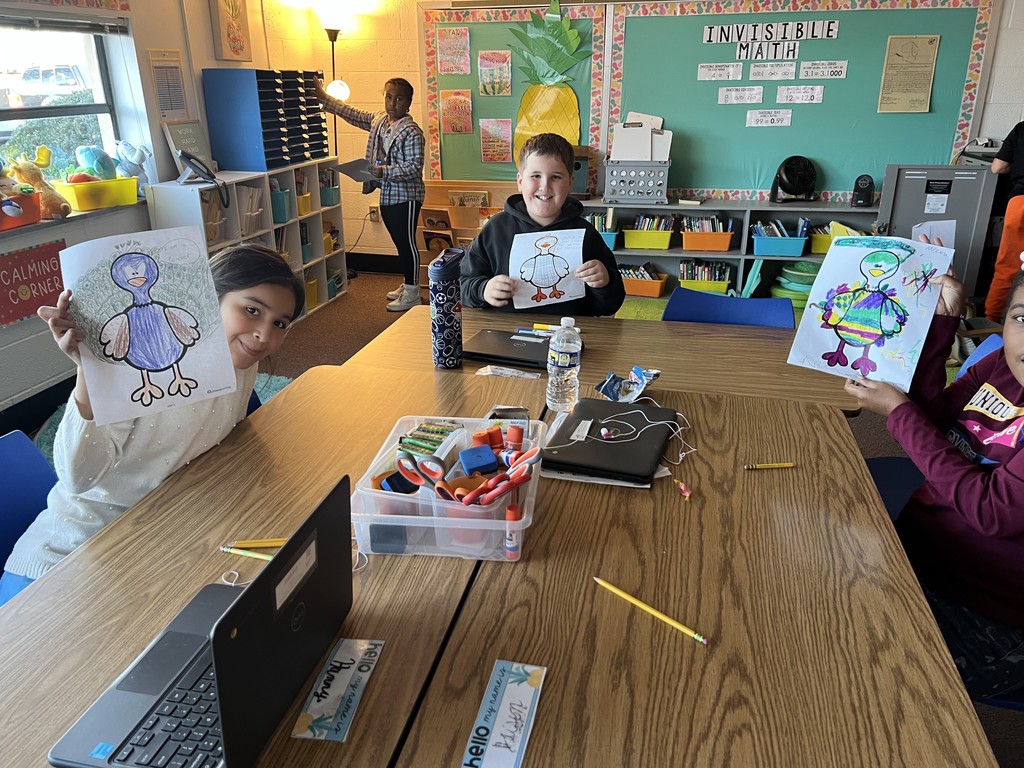 Three students at a table smile while holding up their turkey coloring pages. A fourth student stands at a shelf in the background. The table is covered with pencils, Chromebooks, and a plastic bin of art supplies. The back wall displays an “Invisible Math” bulletin board.