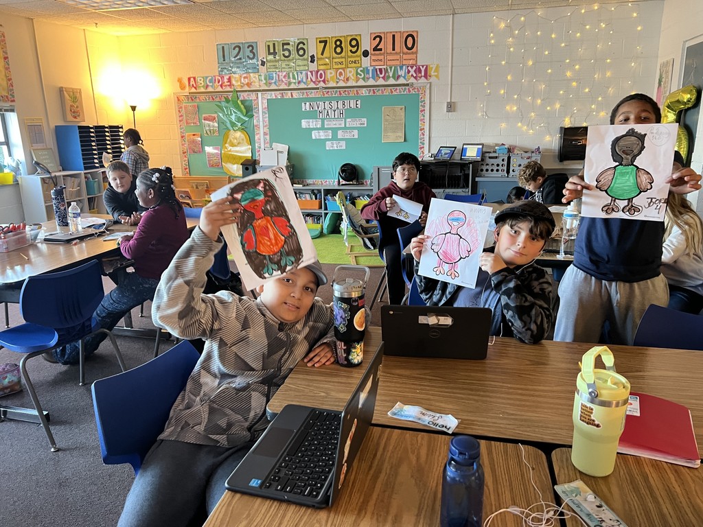 Several elementary students sitting at classroom tables hold up their colored turkey drawings. Laptops, water bottles, and school supplies are on the tables. Other students work in the background under warm lighting and wall decorations with numbers and alphabet letters.