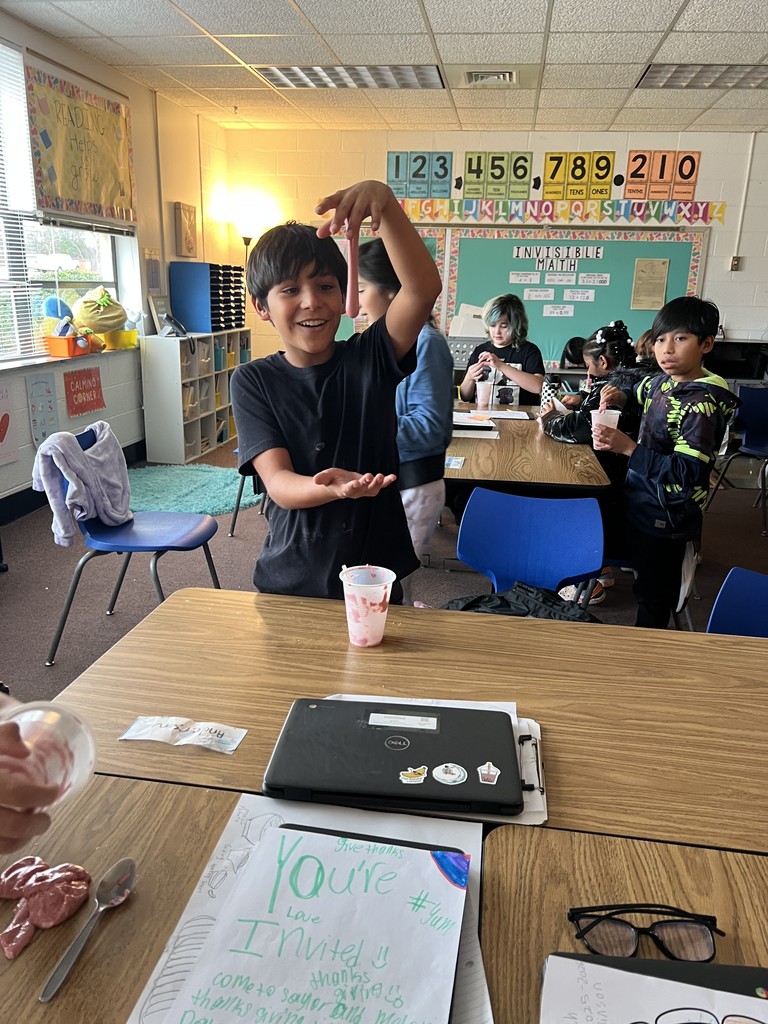 A student stands at a classroom table stretching pink slime between their hands. Other students work and talk in the background. A handwritten paper titled “You’re Invited” is on the table along with a laptop decorated with stickers.