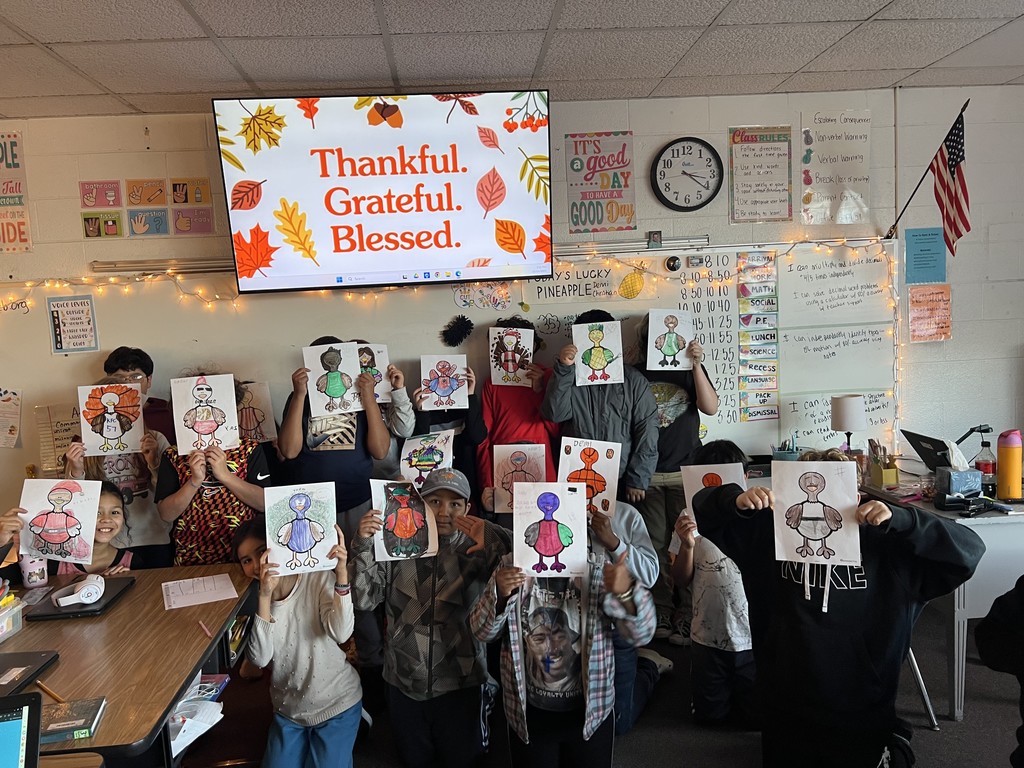 A classroom of elementary students stand and kneel holding up colored turkey drawings in front of their faces. A large screen behind them displays the words “Thankful. Grateful. Blessed.” with fall leaves. The room is decorated with string lights, posters, and classroom charts.