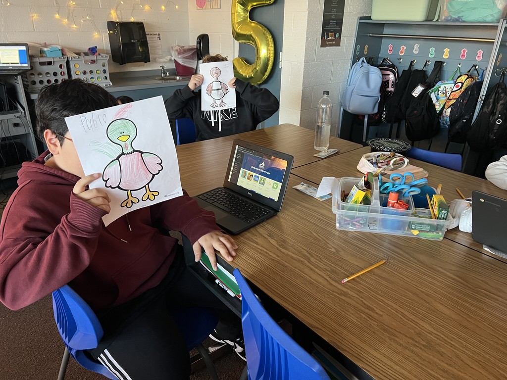 Two students at a classroom table hold up turkey drawings they colored, covering their faces. A laptop and a bin of scissors, crayons, and markers sit on the table. Backpacks hang on hooks along the wall behind them.
