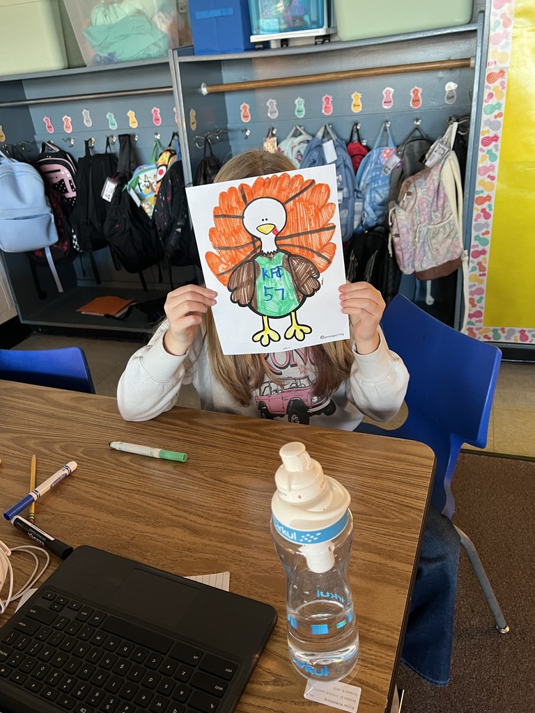 A student sits at a classroom table holding up a brightly colored turkey drawing with orange feathers. A water bottle, Chromebook keyboard, and markers are on the table. Backpacks hang in cubbies behind the student.
