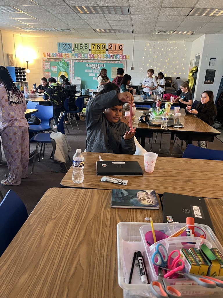 A student sits at a classroom table stretching a long piece of pink slime between their hands. Other students work throughout the room, which is decorated with string lights and colorful bulletin boards. Laptops and school supplies sit on the nearby tables.