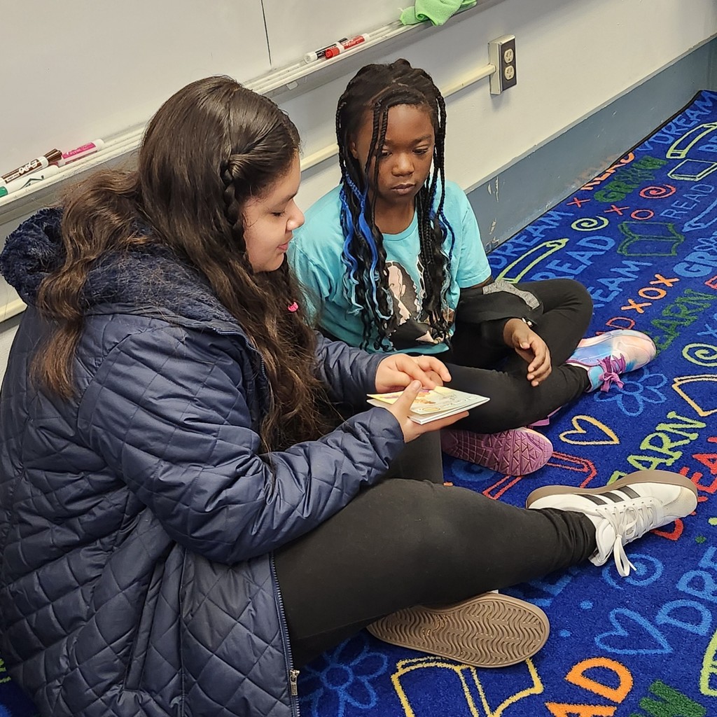 Two students sit together on a colorful classroom rug. One student is holding a book and reading while the other listens closely.
