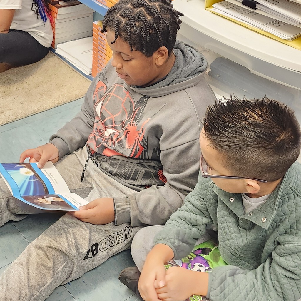 Two young students sit on the carpet looking at an open picture book that shows a space-themed illustration of Earth.