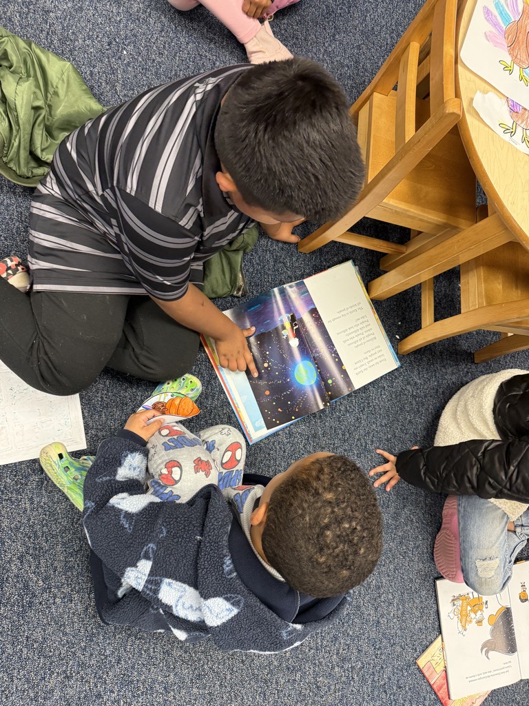 Two students sit on the classroom floor reading a picture book together. One student is holding the book open while the other looks on.