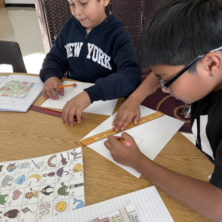 Two students work on a drawing.  They are using a ruler on a wooden table.