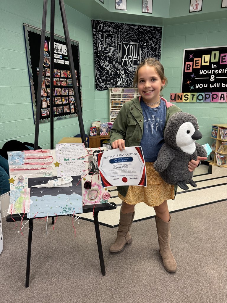 A student smiles while holding a stuffed penguin and a Veterans Day Contest certificate. Beside them, an easel displays their art project with an American flag, watercolor scenes, and handwritten messages thanking veterans. Classroom posters and bookshelves are visible in the background.