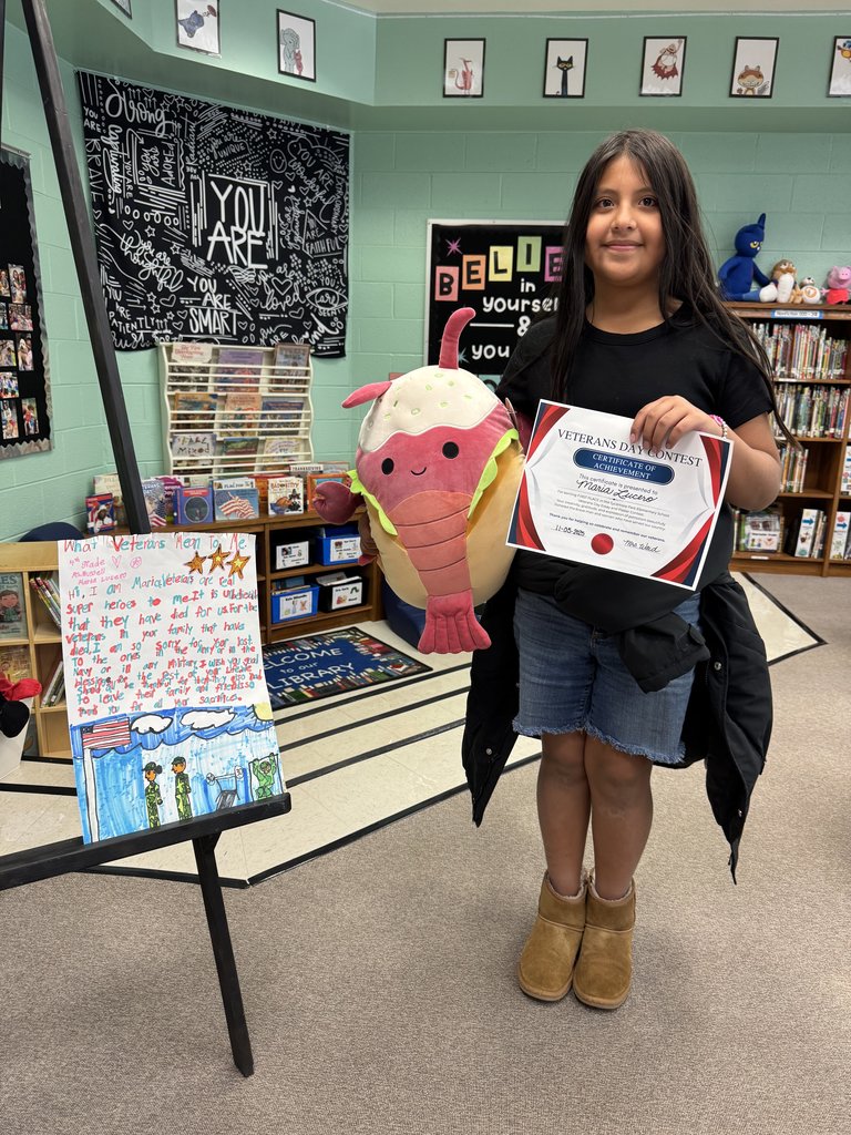 A student holds a large stuffed ice cream plush and a Veterans Day Contest certificate. Their illustrated project on the easel includes colorful drawings of soldiers, clouds, and water, along with a handwritten message about what veterans mean to them.