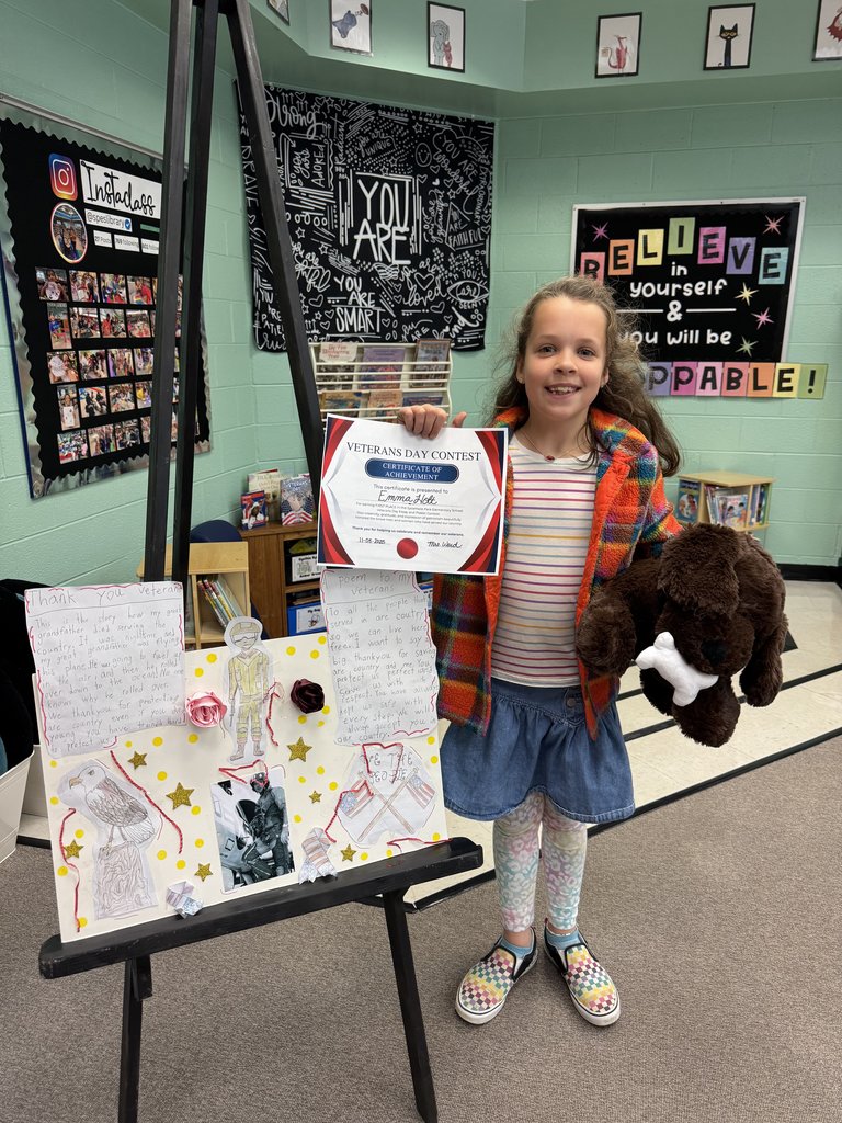 A student stands next to a colorful Veterans Day display board decorated with photos, patriotic symbols, and “Happy Veterans Day! We Thank You.” The student holds a plush dog and a Veterans Day Contest certificate while smiling in a bright library setting.