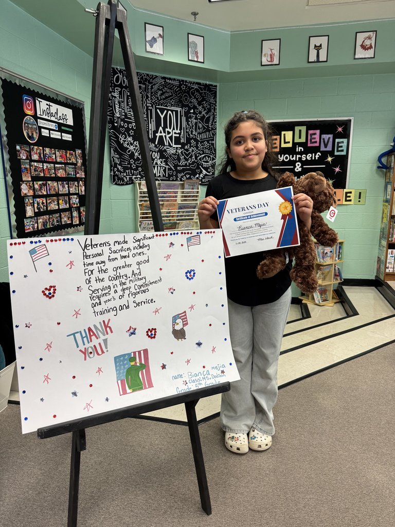 A student holds a plush dog and a Veterans Day Contest certificate. Their large poster displayed on the easel includes handwritten reflections about veterans, patriotic drawings, and decorative stickers. A library space with posters and books is visible behind them.
