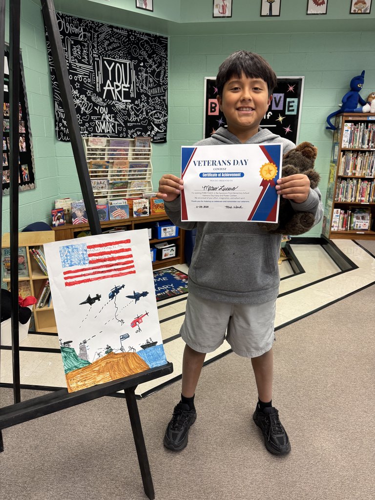 A student in a gray hoodie smiles while holding a Veterans Day Contest certificate and a plush bear. Their artwork on the easel shows a hand-drawn American flag, helicopters, jets, and a landscape scene, all in bright colors.